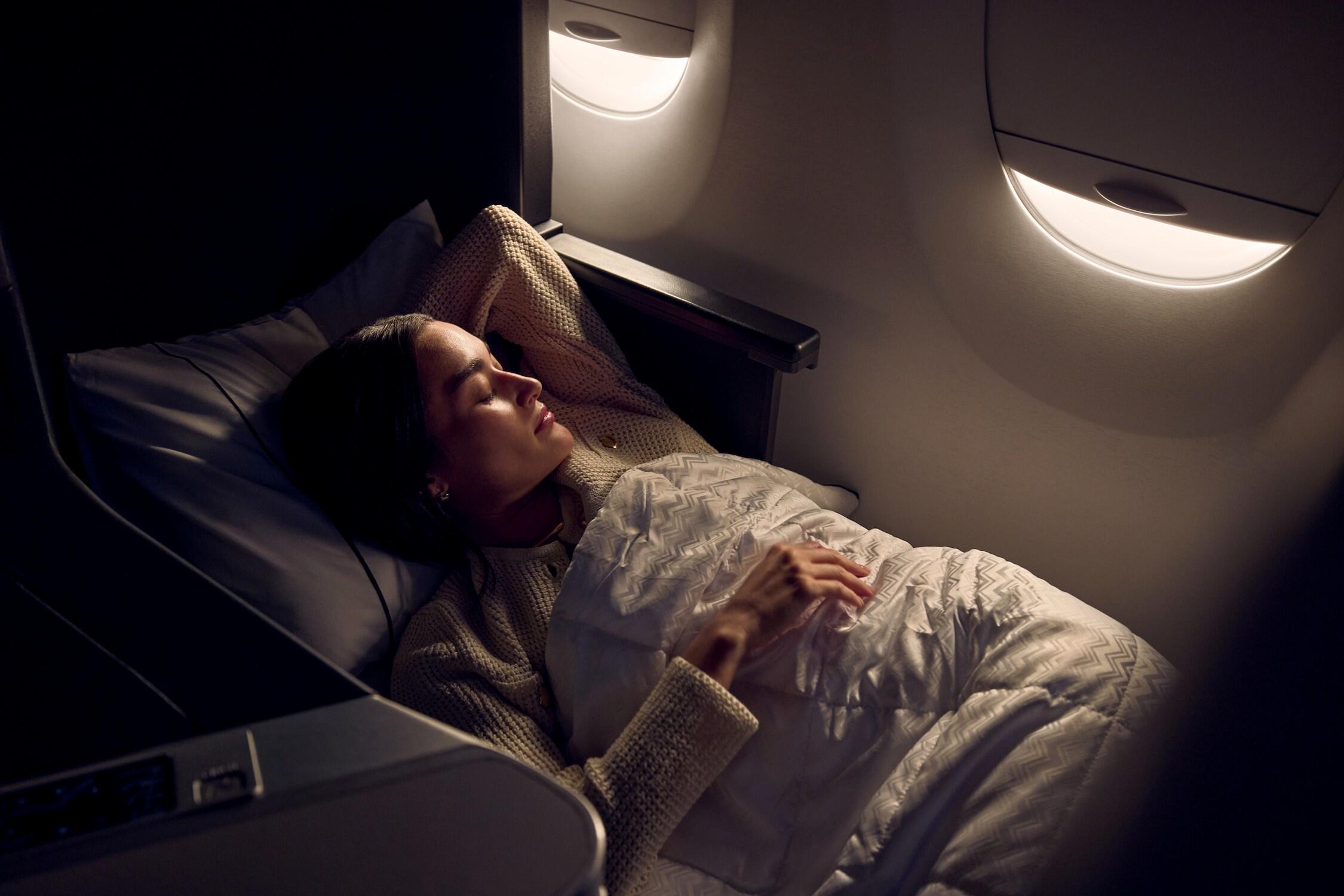 Passenger resting in a lie-flat Delta One seat with bedding, illuminated by soft cabin lighting near an aircraft window.