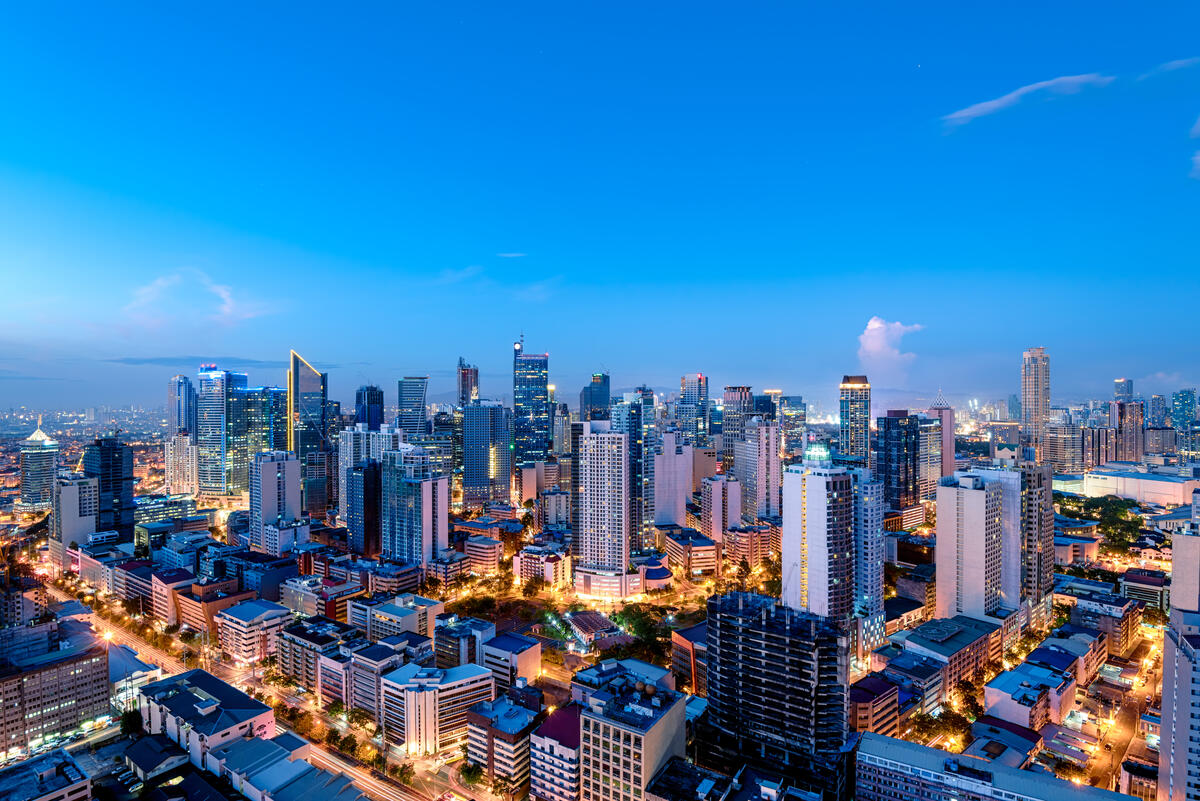 A panoramic view of a modern city skyline at dusk, with illuminated high-rise buildings, busy streets, and a bright blue evening sky.