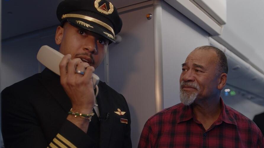 Airline pilot in uniform stands inside an aircraft cabin near the galley, holding an intercom handset. Another person wearing a red plaid shirt is standing beside the pilot.