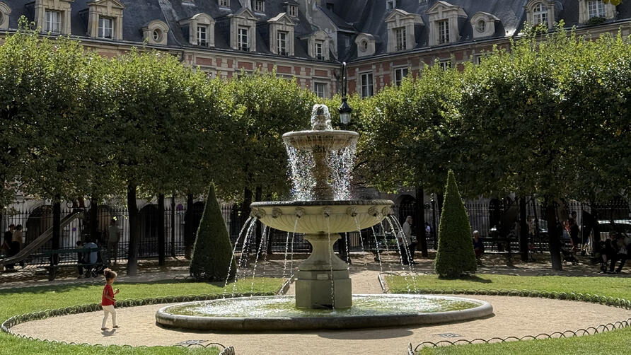 Stone fountain in a leafy Paris square, framed by manicured trees and historic red-brick buildings with slate roofs.