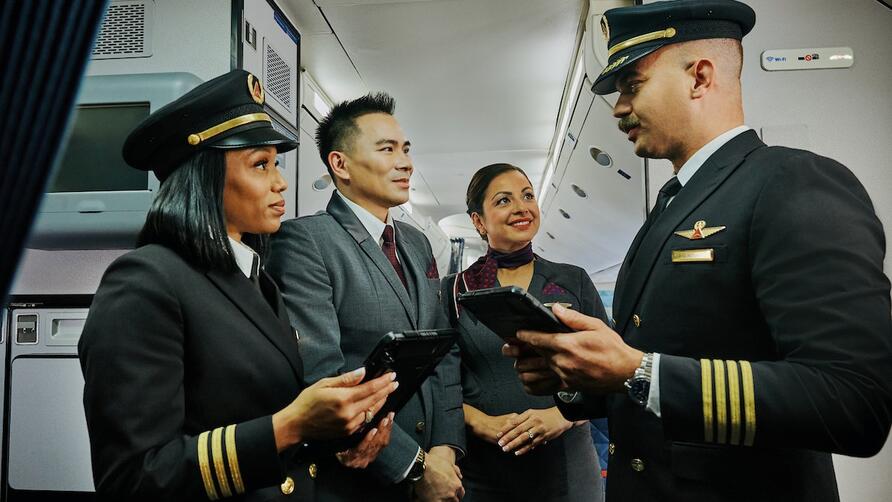 Two Delta pilots and two Delta flight attendants stand together in an airplane aisle, holding tablets and talking.