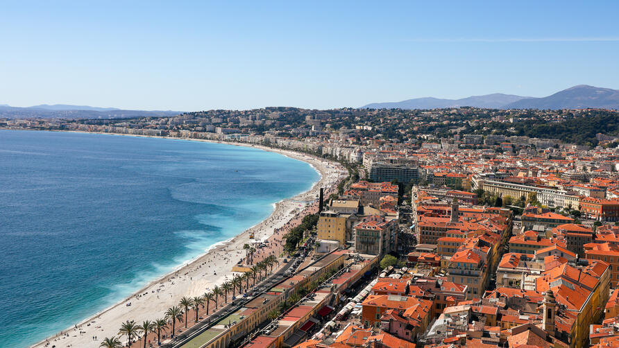Aerial view of a curved Mediterranean beach and waterfront city with red-tiled roofs.