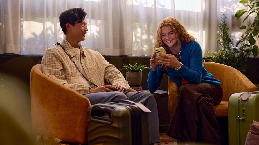 Two travelers sit in lounge chairs with rolling suitcases nearby, one looking at a smartphone, in a softly lit indoor waiting area with plants and sheer curtains.