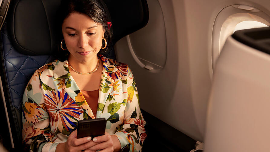 Passenger seated by an airplane window holds a smartphone while sitting in a premium cabin seat, with soft lighting and aircraft interior visible.