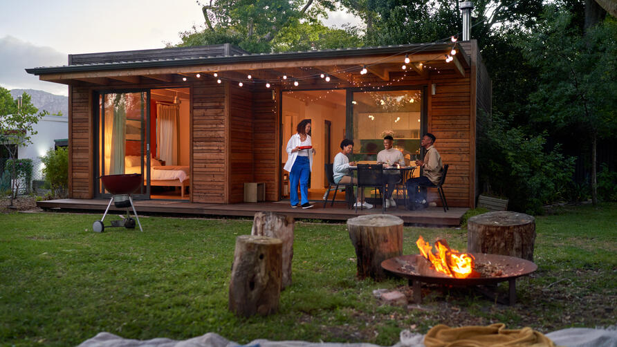 People dining outside a modern wooden cabin at dusk, with string lights, a fire pit and trees surrounding the yard.