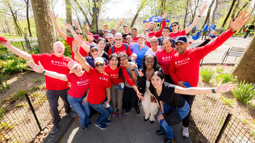 A large group of Delta volunteers wearing red “Delta in the Community” shirts stand together in a park with arms raised, gathered along a paved path surrounded by trees.