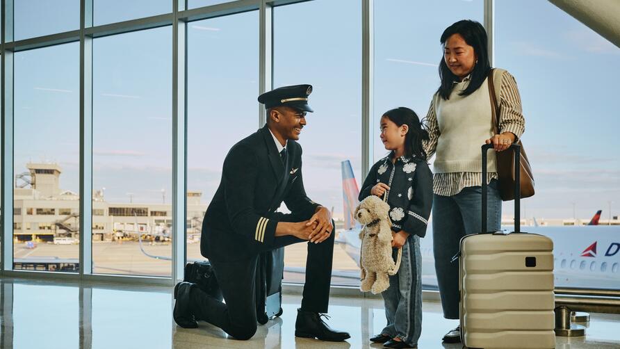 Delta pilot kneeling beside a young child holding a stuffed animal at an airport gate, while an adult stands nearby with rolling luggage; airplane visible through large windows in the background.