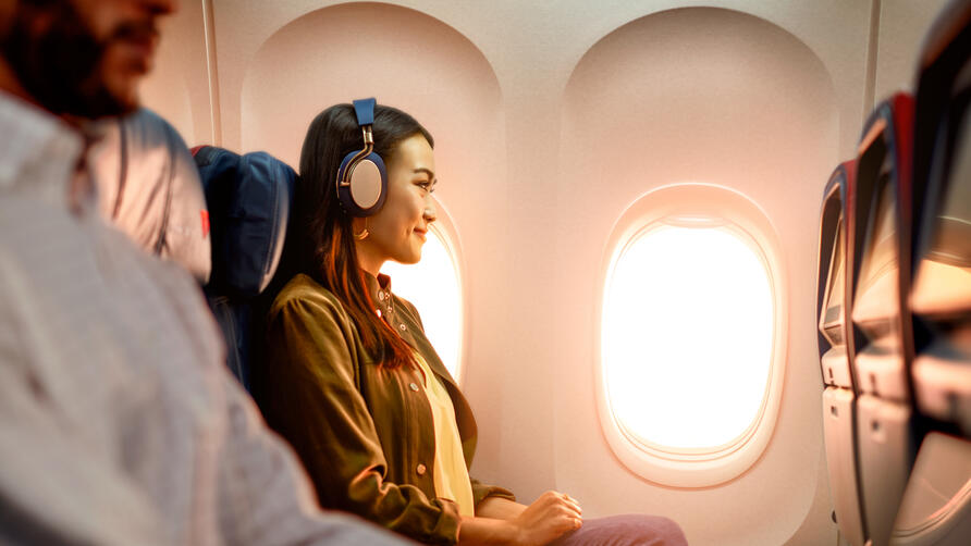 Delta customer on board an airplane wearing headphones sitting by a window, with warm sunlight illuminating the cabin and seatback screens visible.