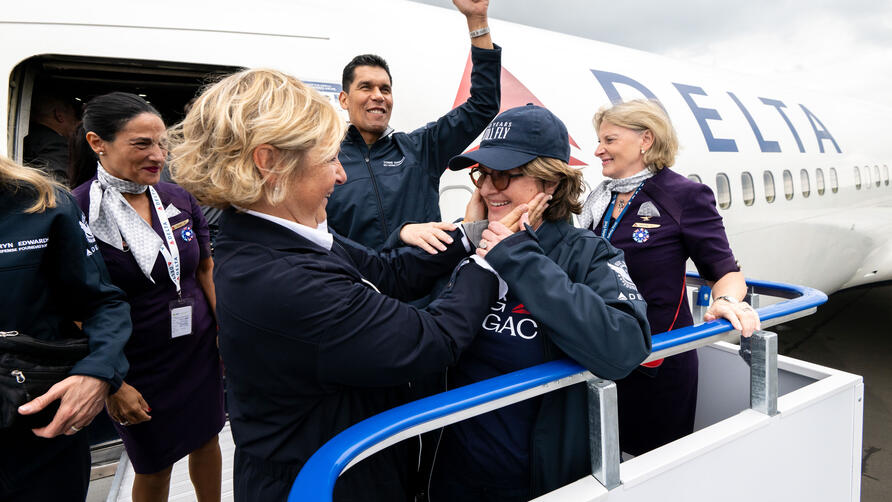 Delta employees and volunteers stand on an airport boarding ramp beside a Delta aircraft, welcoming a passenger as they exit the plane.