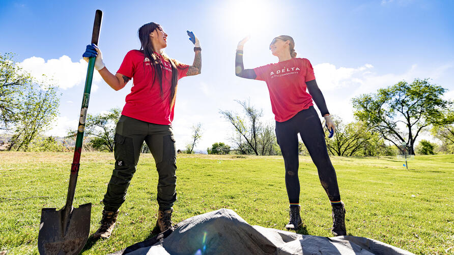 Two volunteers in red “Delta in the Community” shirts high-five while working outdoors, one holding a shovel on a sunny grassy field with trees in the background.