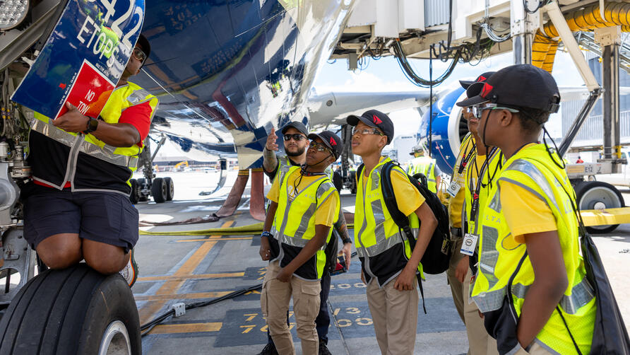 Students in safety vests gather beneath a Delta aircraft as an aviation professional points out features near the landing gear on the airport ramp.