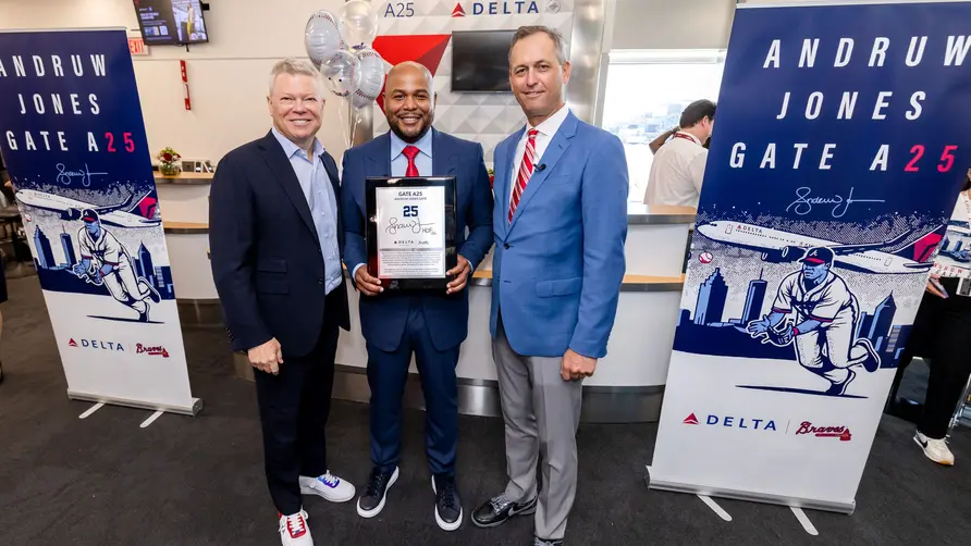 Three people pose at a Delta airport gate ceremony, with the center person holding a commemorative plaque. Banners on both sides read “Andruw Jones Gate A25,” featuring baseball-themed artwork and Delta branding.