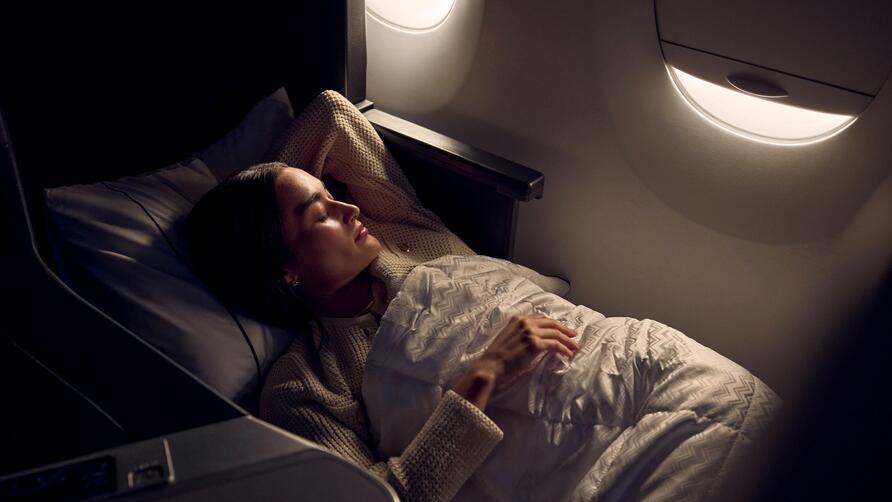  Passenger resting in a lie-flat Delta One seat with bedding, illuminated by soft cabin lighting near an aircraft window.