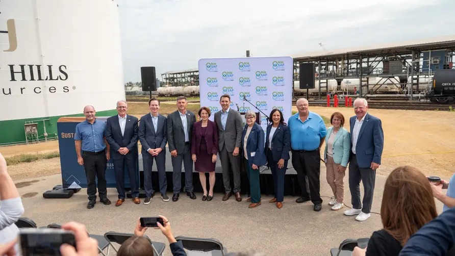 Group photo of event attendees standing in front of an industrial biofuels facility, with storage tanks, railcars, and a branded backdrop visible behind them while audience members take photos in the foreground.