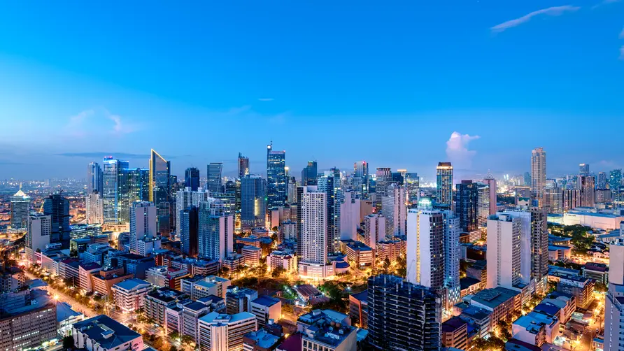 A panoramic view of a modern city skyline at dusk, with illuminated high-rise buildings, busy streets, and a bright blue evening sky.