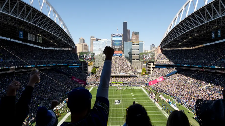 Fans in a packed football stadium watch the game from the stands, with the field and Seattle skyline visible beyond the open end of the venue.