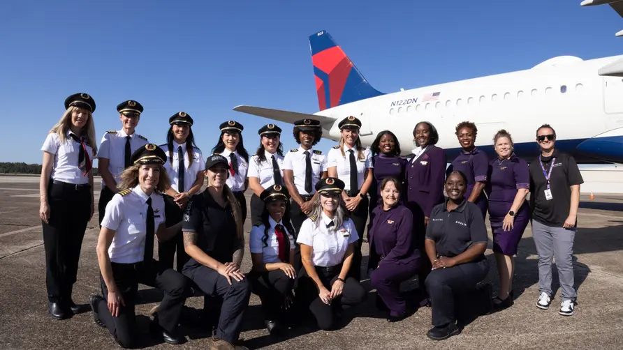 Group of aviation professionals and participants posing on the tarmac in front of a Delta aircraft under a clear blue sky during a Girls in Aviation Day event.