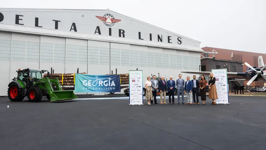 Group stands in front of Delta Air Lines’ historic hangar at the Delta Flight Museum alongside a tractor, timber load and Georgia SAF Coalition signage during a sustainable aviation fuel event.