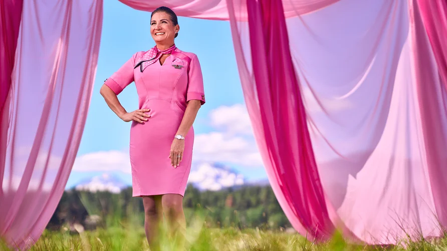 Person in a pink uniform dress standing outdoors between flowing pink fabric panels, with grass, trees and mountains in the background.
