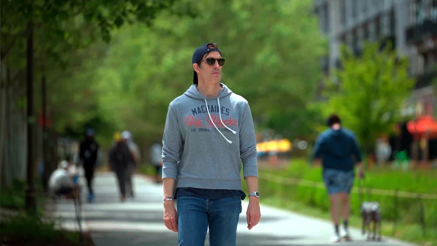 Person wearing a gray hoodie, sunglasses and a backward cap walking along a tree-lined urban pathway, with other pedestrians in the background.