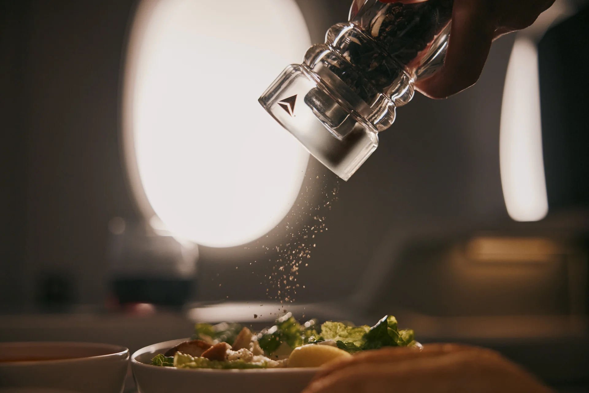Close-up of a pepper grinder seasoning a fresh salad during an in-flight dining service, with airplane windows softly lit in the background.