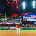 Fireworks light up a baseball stadium at night as two players stand together on the infield, with a scoreboard displaying “Braves Win” and a packed crowd in the background.