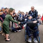 A seated veteran in a wheelchair shakes hands with a child in a green uniform as a crowd lines the roadway, applauding during a commemorative event.