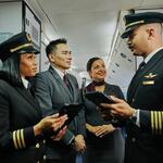 Two Delta pilots and two Delta flight attendants stand together in an airplane aisle, holding tablets and talking.