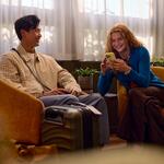 Two travelers sit in lounge chairs with rolling suitcases nearby, one looking at a smartphone, in a softly lit indoor waiting area with plants and sheer curtains.