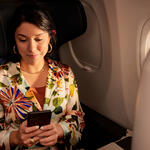 Passenger seated by an airplane window holds a smartphone while sitting in a premium cabin seat, with soft lighting and aircraft interior visible.