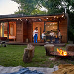 People dining outside a modern wooden cabin at dusk, with string lights, a fire pit and trees surrounding the yard.