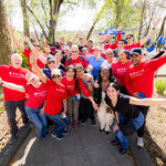 A large group of Delta volunteers wearing red “Delta in the Community” shirts stand together in a park with arms raised, gathered along a paved path surrounded by trees.