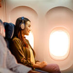 Delta customer on board an airplane wearing headphones sitting by a window, with warm sunlight illuminating the cabin and seatback screens visible.