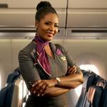 Flight attendant standing in an airplane cabin aisle, wearing a uniform with a neck scarf, arms crossed, with passenger seats and windows in the background.