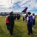 Spectators stand on grass watching a Delta airplane taxi on the runway at an airport.