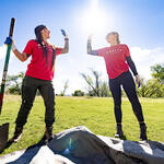 Two volunteers in red “Delta in the Community” shirts high-five while working outdoors, one holding a shovel on a sunny grassy field with trees in the background.