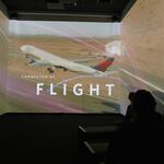 Silhouetted audience in a dark room viewing a multi-wall projection of a plane taking off, with the text “CONNECTED BY FLIGHT” overlaid.