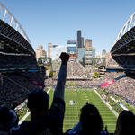 Fans in a packed football stadium watch the game from the stands, with the field and Seattle skyline visible beyond the open end of the venue.