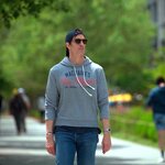 Person wearing a gray hoodie, sunglasses and a backward cap walking along a tree-lined urban pathway, with other pedestrians in the background.