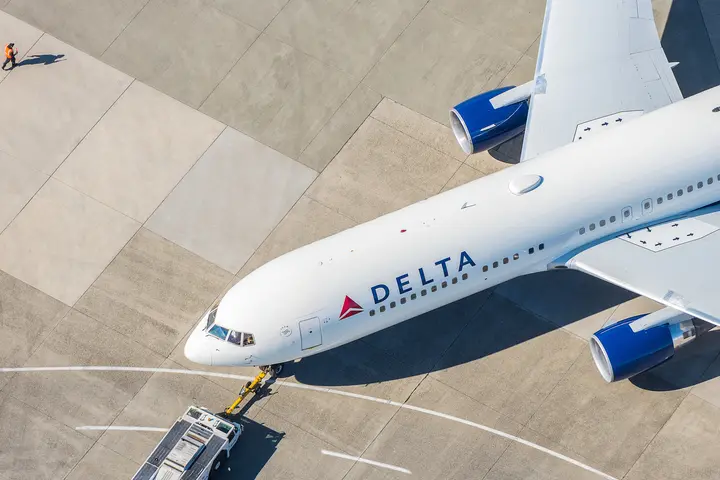 A 767-300 on the runway complemented by various objects in the background (boundary lines on the runway & a below wing employee).