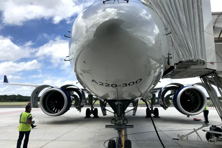 Nose of A220-300 parked on tarmac with stair truck and employees