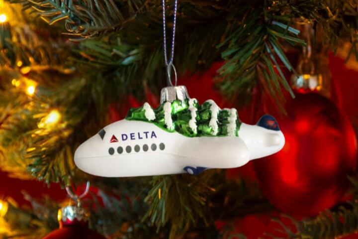 Soar into style with our Festive Delta Plane Ornament. Made of glass with a whimsical tree on top, make sure to snag one before it takes off! 