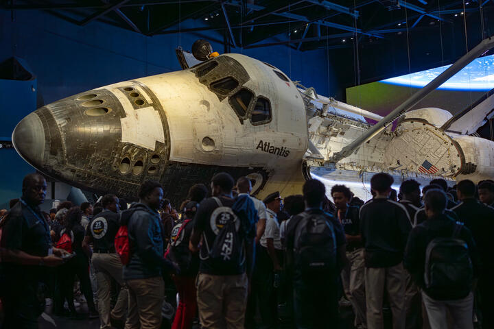 Space Shuttle Atlantis displayed in a museum exhibit with DREAM flight attendees gathered around.