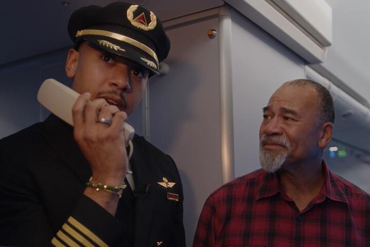 Airline pilot in uniform stands inside an aircraft cabin near the galley, holding an intercom handset. Another person wearing a red plaid shirt is standing beside the pilot.