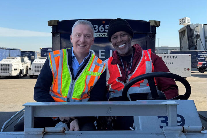 Two workers in high-visibility vests sitting on an airport ground vehicle near Delta equipment and a boarding gate.