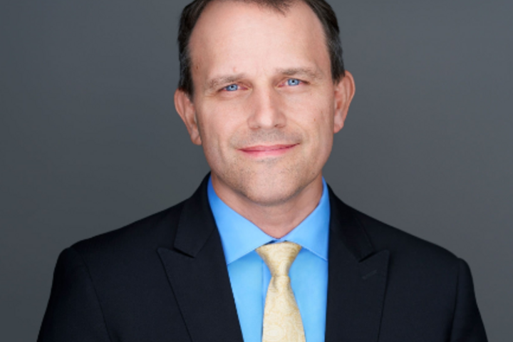 Headshot of Matt Schrag, wearing a dark suit jacket, light blue dress shirt and light yellow tie, posed against a plain gray background.