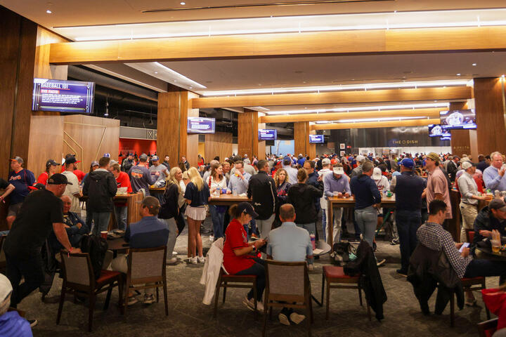 A crowded indoor stadium lounge with fans standing and sitting at tables beneath wood-paneled ceilings and multiple TV screens.