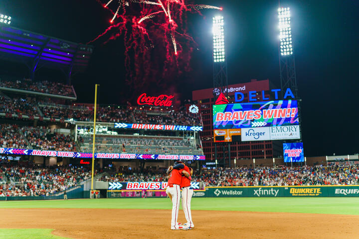 Fireworks light up a baseball stadium at night as two players stand together on the infield, with a scoreboard displaying “Braves Win” and a packed crowd in the background.