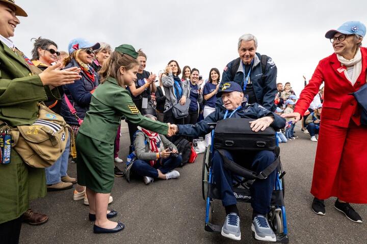 A seated veteran in a wheelchair shakes hands with a child in a green uniform as a crowd lines the roadway, applauding during a commemorative event.