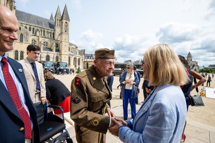 A uniformed World War II–era veteran shakes hands with an attendee in front of a historic stone church, with a small group of people gathered nearby at an outdoor commemorative event.